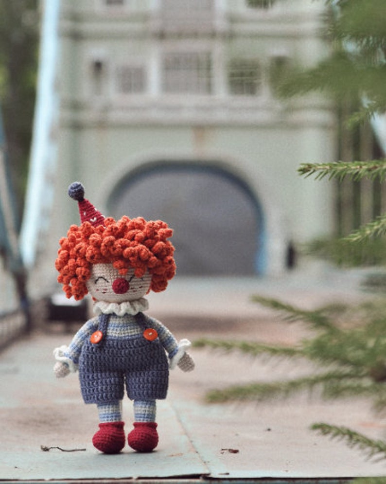 May include: A crocheted clown doll with red hair, a blue and white striped outfit, and a red nose. The doll is standing on a gray surface.
