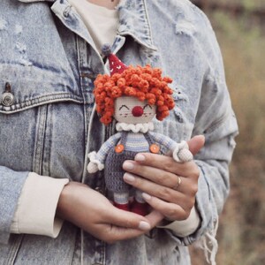 May include: A crocheted clown doll with red hair, a white collar, and blue and white striped overalls. The doll is being held by a person wearing a denim jacket.