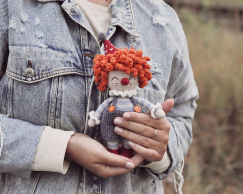 May include: A crocheted clown doll with red hair, a white collar, and blue and white striped overalls. The doll is being held by a person wearing a denim jacket.