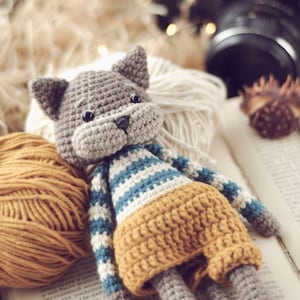 Op de afbeelding: Een gehaakte grijze kat die een blauw-wit gestreept shirt en een gele broek draagt. De kat zit op een boek met garenbollen en een camera op de achtergrond.