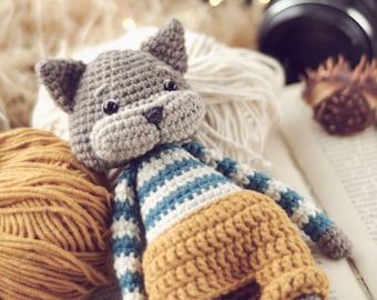 May include: A crocheted gray cat wearing a blue and white striped shirt and yellow shorts. The cat is sitting on a book with yarn balls and a camera in the background.