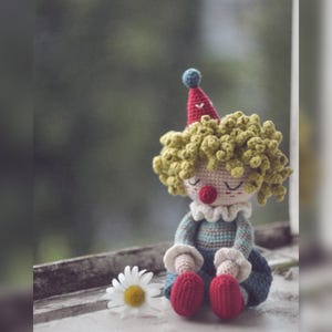 Puede incluir: Una muñeca payaso de ganchillo con pelo rizado verde claro, nariz roja y un sombrero cónico rojo con un pompón azul. La muñeca lleva una camiseta de manga larga a rayas azules y verdes con puños y cuello de volantes blancos, y zapatos rojos. Una margarita blanca se encuentra al lado.
