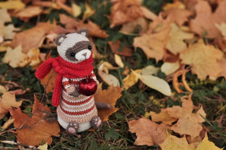 Puede incluir: Un juguete de mapache de crochet que lleva una bufanda roja y sostiene una manzana roja. El juguete est&aacute; sentado en una cama de hojas de oto&ntilde;o.