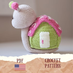 Puede incluir: Un patrón de ganchillo en PDF de un juguete con forma de caracol-casa. El caracol tiene cabeza y cuerpo de color beige claro, dos pequeños pompones en la cabeza y una concha con forma de casa con paredes verdes, techo rosa y puerta blanca.