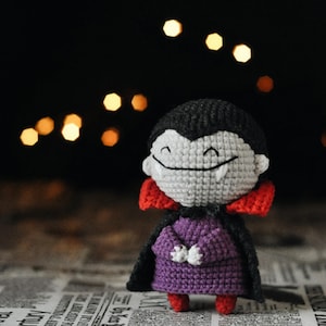 Puede incluir: Una muñeca de vampiro tejida a ganchillo con una cabeza gris grande, cabello negro y una amplia sonrisa que muestra dos colmillos blancos. La muñeca viste una túnica morada, una capa negra con forro rojo y zapatos rojos pequeños.