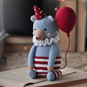 Puede incluir: Un oso de crochet azul con un atuendo a rayas rojas y blancas y un sombrero de lunares rojos y blancos. El oso sostiene un globo rojo.