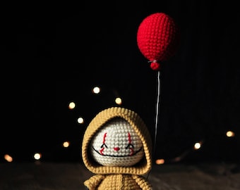 Könnte beinhalten: Eine gehäkelte Puppe, die einen gelben Regenmantel trägt und einen roten Ballon hält. Die Puppe hat ein weißes Gesicht mit schwarzen Augen und einem roten Lächeln.