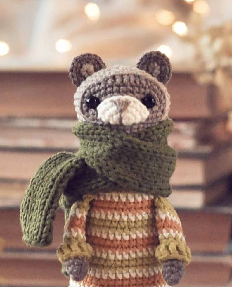 Puede incluir: Un animal de peluche tejido a crochet con una bufanda verde y un su&eacute;ter a rayas. El animal tiene una cara marr&oacute;n y gris con ojos negros y una nariz blanca.
