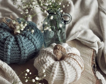 May include: Two crocheted pumpkins, one teal and one white, sit on a wooden slice with a vase of white baby's breath.