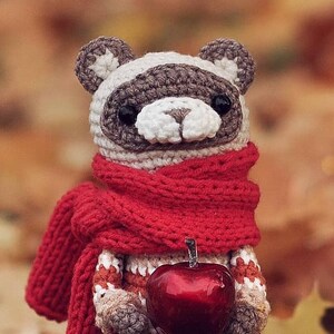 Puede incluir: Un animal de peluche tejido a crochet que lleva una bufanda roja y sostiene una manzana roja. El animal tiene una cara gris y blanca con ojos negros.