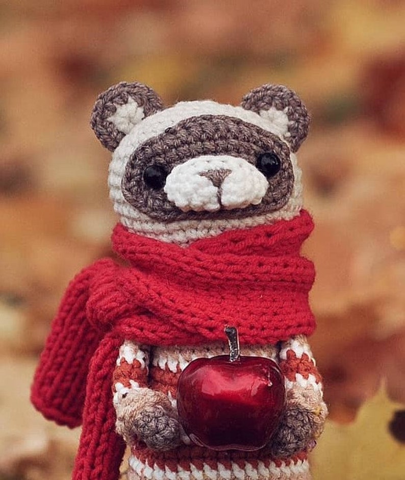 Puede incluir: Un animal de peluche tejido a crochet que lleva una bufanda roja y sostiene una manzana roja. El animal tiene una cara gris y blanca con ojos negros.