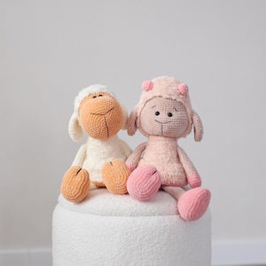 Può includere: Due bambole di pecore fatte a mano all'uncinetto. Una è bianca con viso color crema e piedi arancioni, l'altra è rosa con cappello abbinato e piedi rosa. Entrambe le bambole sono sedute su una superficie bianca strutturata.