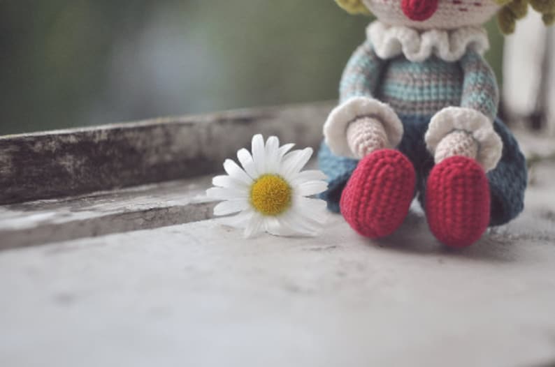 May include: A crocheted doll with a red nose and red shoes sits on a windowsill with a white daisy in front of it.