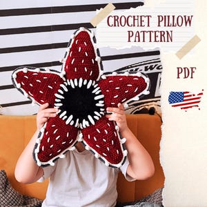 Può includere: Un cuscino all'uncinetto a forma di stella, color bordeaux con dettagli bianchi e un centro nero. Il cuscino è tenuto da una persona. Il testo "CROCHET PILLOW PATTERN" è visibile.