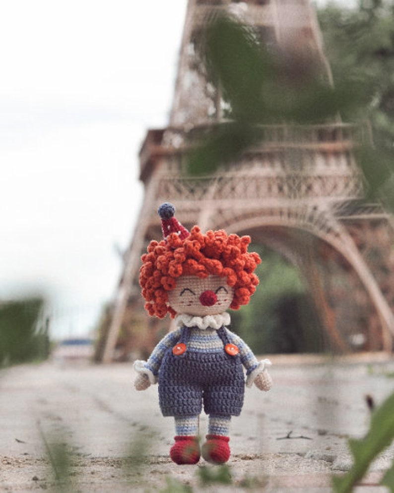 May include: A crocheted clown doll with red hair, a blue and white striped outfit, and a red nose. The doll is standing in front of a blurred background of a building.