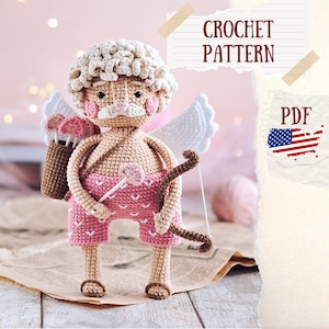 Puede incluir: Patrón de crochet PDF para un amigurumi de crochet de Cupido. La muñeca lleva pantalones cortos rosas con corazones blancos, tiene un carcaj marrón con flechas y sostiene una flecha rosa en forma de corazón. La muñeca tiene un halo blanco de crochet y un bigote marrón.