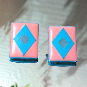 May include: Two small, rectangular hair clips. Each clip has a pink background with a blue diamond shape in the center. The clips have a blue base and are displayed on a reflective surface.
