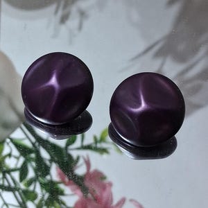 May include: Two round, dark purple buttons with a starburst design on a reflective surface. The buttons have a glossy finish and are slightly raised. The background includes greenery and pink flowers, adding a touch of color.