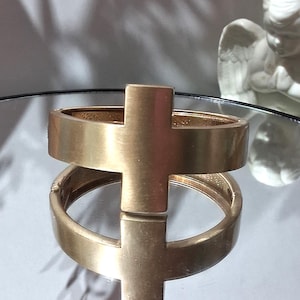 May include: A gold-colored bracelet with a cross design. The bracelet is displayed on a reflective surface, with a small statue of an angel in the background. The cross is centered on the bracelet.