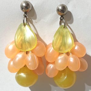 Puede incluir: Un par de pendientes vintage con un diseño de racimo. Cada pendiente tiene un perno de color plateado, un acento en forma de lágrima amarilla y un grupo de cuentas de color melocotón. La cuenta inferior es amarilla.