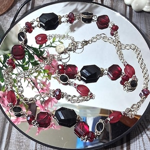 May include: A silver-toned necklace with alternating black and red faceted beads. The necklace features a chain link design and is displayed on a reflective surface. The beads are various shapes and sizes.