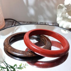 May include: Two wooden bangles, one a rich reddish-brown and the other a darker, patterned brown. The bangles are displayed on a reflective surface, creating a mirrored effect. The background includes a small statue and floral accents.