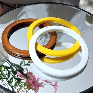 May include: Three circular bag handles in different colors: a dark brown wood, a bright yellow, and a white. The handles are arranged on a reflective surface, with a hint of greenery and pink flowers in the background.