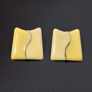 May include: A pair of square-shaped stud earrings featuring a smooth, glossy finish. Each earring is divided into two sections, one light yellow and one cream, separated by a thin, vertical, wavy silver-toned metal line through the center.