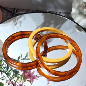 May include: Four vintage bangles in shades of amber and cream are arranged on a reflective surface. The bangles have a smooth, glossy finish, with the amber ones displaying a marbled effect. The jewelry is a classic accessory.