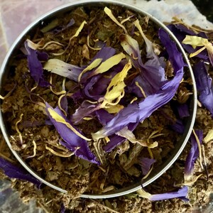 May include: A close-up of a metal container filled with dried herbs and flower petals. The herbs are a dark brown color, and the petals are a mix of purple and yellow.