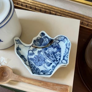 May include: A small, blue and white porcelain teapot-shaped dish with floral patterns. The dish is placed on a stack of books, with a wooden spoon and a white ceramic container with a blue rim in the background. The spoon has the word "PORTUGAL" engraved on it.