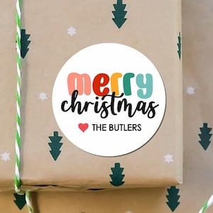 May include: A wrapped gift with a round sticker that says "Merry Christmas" in colorful letters. The sticker also includes a red heart and the name "THE BUTLERS". The wrapping paper is tan with green Christmas trees and white stars.