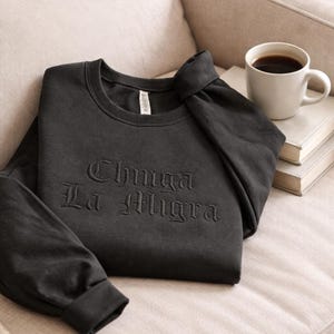 May include: A dark gray sweatshirt with the embroidered text "Chinga La Migra" is folded on a beige sofa. A white mug filled with coffee sits on a stack of books next to the sweatshirt. The sweatshirt has long sleeves and a crew neck.