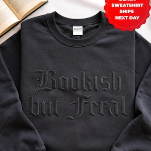 May include: Black sweatshirt with the embroidered phrase "Bookish but Feral" in gothic lettering. The crew neck sweatshirt is a dark color and has a tag that says "Gildan". A red tag with white text says "Black Sweatshirt Ships Next Day".