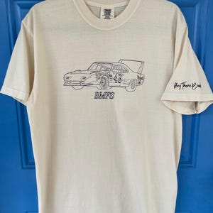 May include: Cream-colored t-shirt featuring a black line drawing of a vintage race car with the number 43. The text "BMFS" is below the car. The sleeve has the text "Hey There Bub".