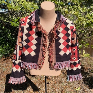 May include: A black, red, white, and brown patterned cardigan with fringe trim. The cardigan has a geometric design and is made of a soft, woven fabric.