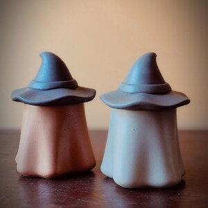 Peut inclure: Deux figurines en céramique en forme de sorcière. L'une est de couleur pêche, l'autre est blanc cassé. Les deux ont des chapeaux de sorcière gris. Les figurines sont sur une surface marron foncé.