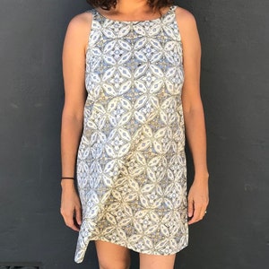May include: A woman wearing a sleeveless dress with a blue and white geometric pattern. She is wearing a straw hat and black flip flops.