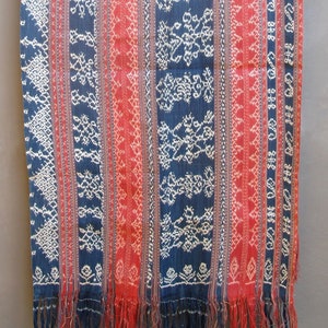 May include: A textile with a striped pattern in shades of blue and red, with white embroidered details. The fabric has a fringed edge and is hanging from a wooden rod. The design features geometric and floral motifs.
