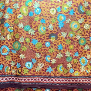 May include: Close-up of a textile with an intricate floral pattern. The design features teal, green, and gold flowers and leaves on a burgundy background. A decorative border with a repeating pattern is visible.