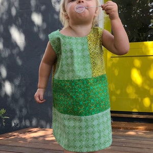 May include: A young child wearing a green and yellow patchwork dress with a white geometric pattern. The dress has a sleeveless design and a scoop neck. The child is standing on a wooden deck and blowing bubbles.