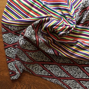 May include: A folded textile with vibrant, multicolored stripes and a patterned border. The stripes include red, orange, yellow, green, blue, and purple. The border features a geometric diamond pattern in black, white, and red.