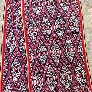 May include: A long, rectangular scarf with a repeating diamond pattern in red, white, and black. The edges are trimmed with yellow and red, and the ends have a fringe. The scarf is made of a woven fabric.