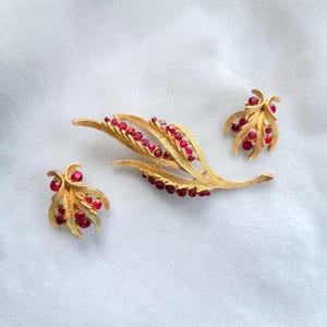 May include: A gold-tone jewelry set consisting of a leaf-shaped brooch and two matching earrings. Each piece features a textured, brushed gold finish and is accented with small, round, deep red faceted stones arranged in a cluster or line pattern.