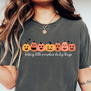 May include: A dark grey t-shirt with a line of orange and brown jack-o'-lantern pumpkins with different facial expressions. The text "helping little pumpkins do big things" is printed below the pumpkins.