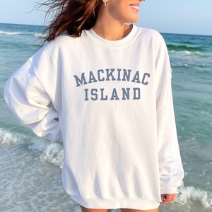 May include: White sweatshirt with blue text that reads "MACKINAC ISLAND" on the front. The sweatshirt is worn by a person standing on a beach.