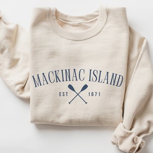 May include: A beige sweatshirt with the text "MACKINAC ISLAND EST 1671" and a pair of crossed oars in navy blue.