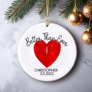 May include: White ceramic Christmas ornament with a red heart design and the words "Better Than Ever". The heart has a stitched design. The ornament also includes the name "Christopher" and the date "3.5.2022".