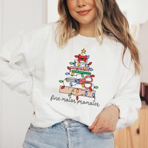 May include: White sweatshirt featuring a Christmas tree design made from school supplies, including scissors, pencils, a ruler, and a pencil sharpener. The text "fine motor promoter" is written below the tree.
