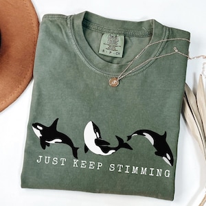 Op de afbeelding: Olijfgroen t-shirt met een afbeelding van drie zwart-witte orka's en de tekst "JUST KEEP STIMMING" in het wit. Een gouden ketting met een hanger ligt op het shirt.